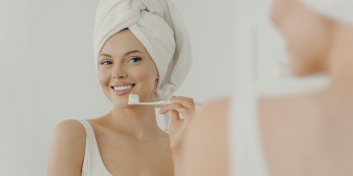 Beautiful young woman with healthy perfect smile brushing teeth and looking in mirror